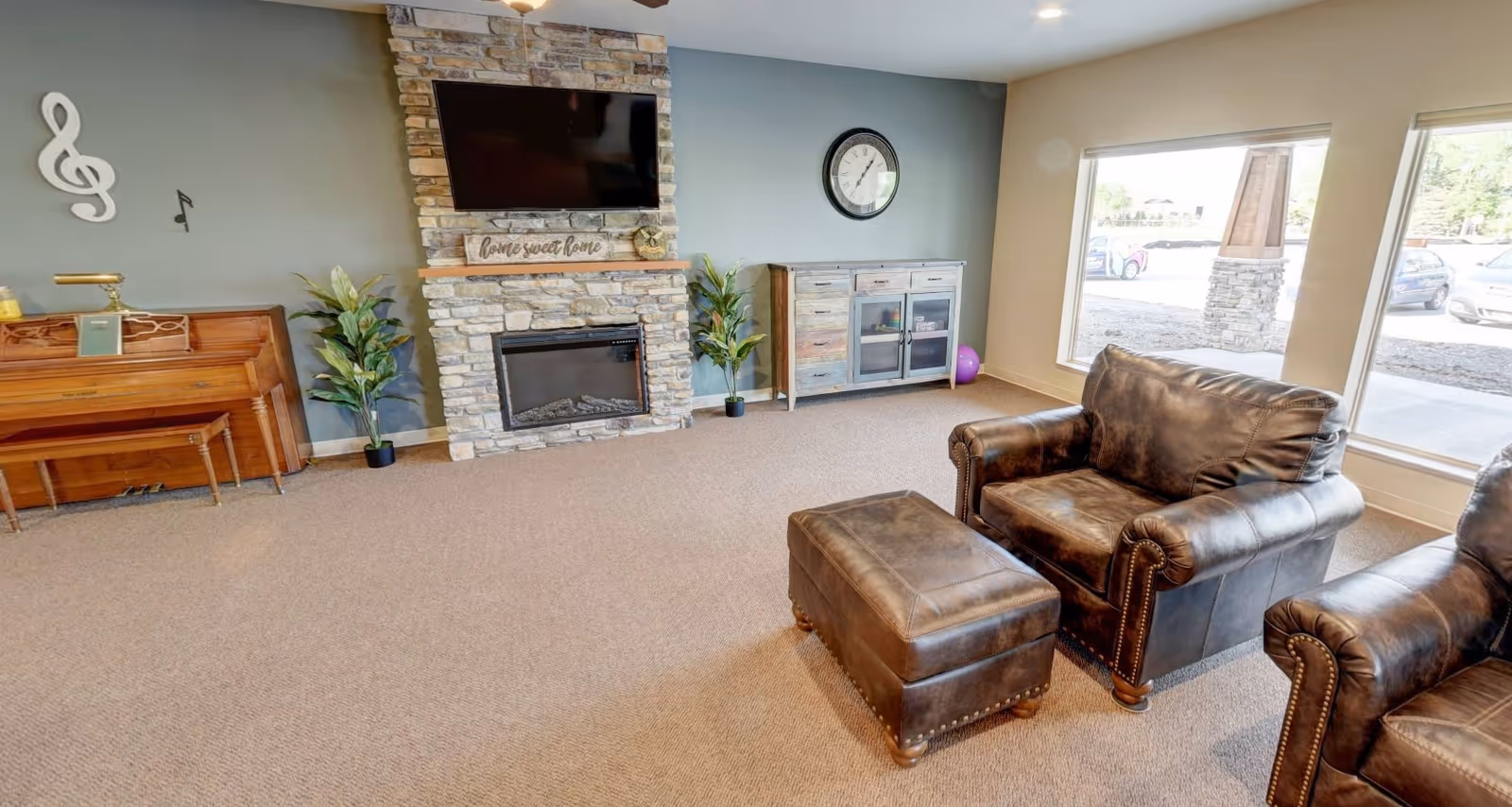 Spacious common living room with a stone fireplace and wall-mounted TV, piano, leather armchairs with ottoman, and large windows.
