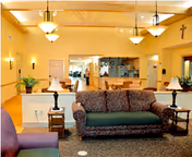 A spacious living room area with a patterned sofa, two side tables with lamps, and an armchair. The room features warm yellow walls, wooden ceiling beams, and hanging light fixtures. In the background, there is a kitchen area with counters and stools, and a cross mounted on the wall to the right.