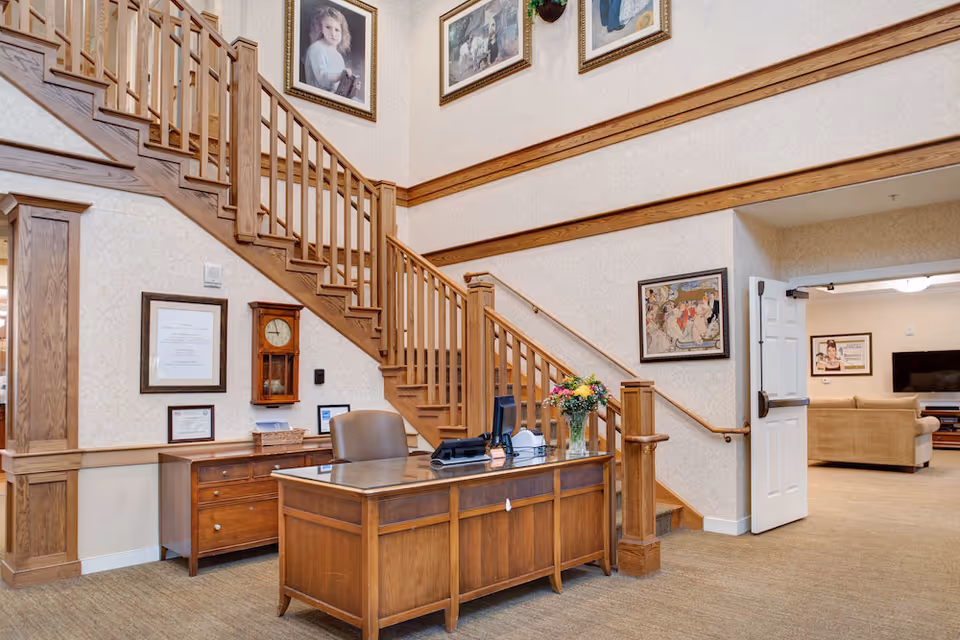 Reception desk and wooden staircase in a senior living facility lobby with framed artwork and seating visible through a doorway.