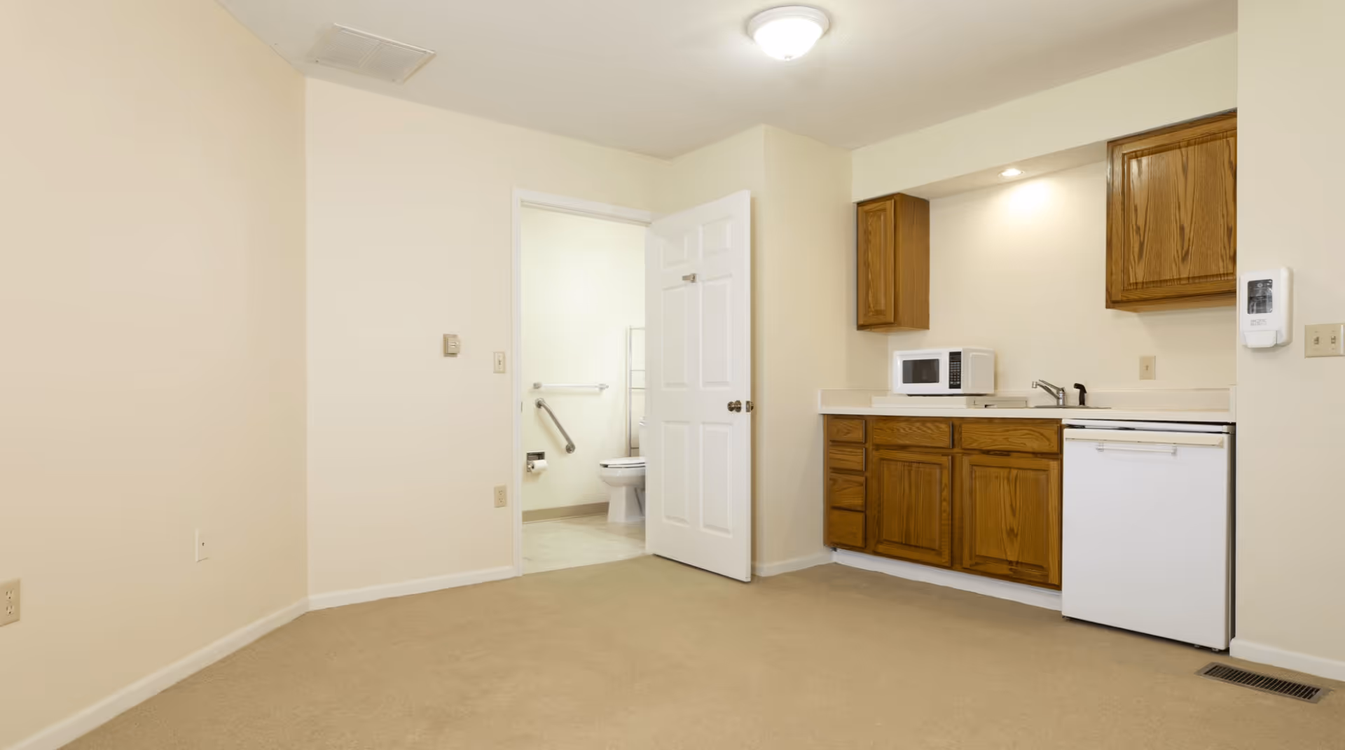 Small assisted-living studio interior with a kitchenette (wood cabinets, microwave, mini-fridge) and an open door revealing a bathroom with a toilet and grab bar.