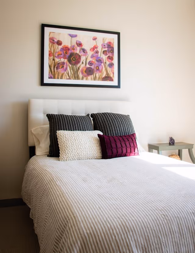 A neatly made bed with striped bedding and decorative pillows beneath a framed floral painting in a bright bedroom.