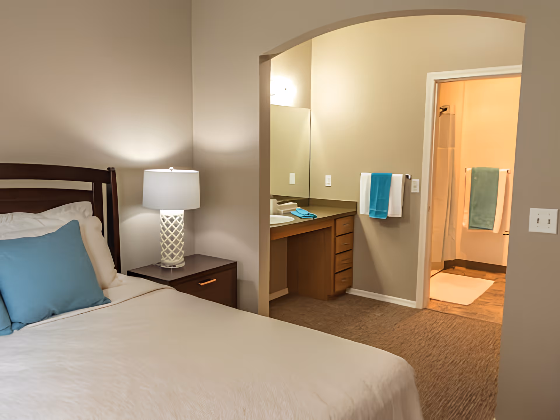 A bedroom with a neatly made bed featuring white bedding and blue pillows. Next to the bed is a dark wooden nightstand with a white lamp. The room has beige walls and carpeted floor. An open doorway leads to a bathroom area with a vanity, mirror, and towels hanging on the wall. The bathroom has a shower with a curtain and additional towels.