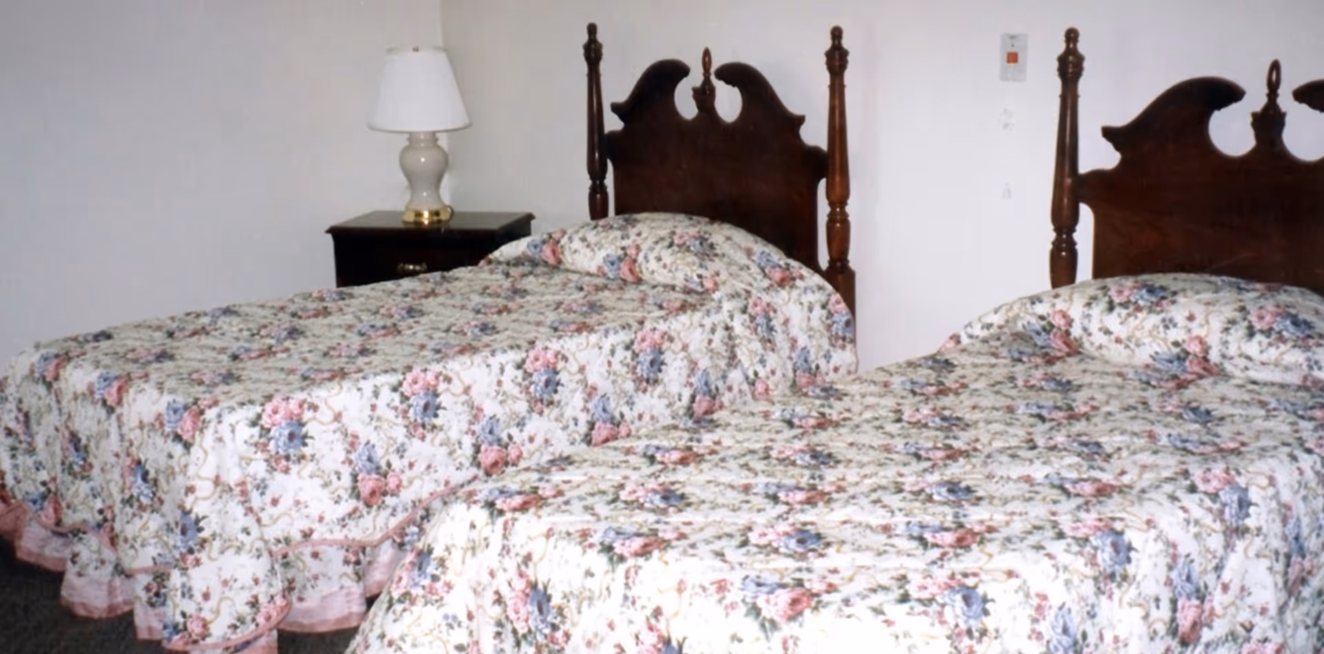 Two twin beds with floral bedspreads and carved wooden headboards flanking a nightstand with a lamp in a simple bedroom.