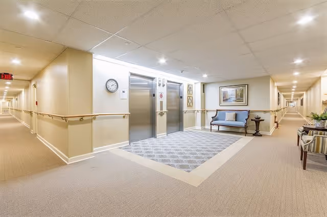 Well-lit hallway in a senior living facility with two stainless steel elevators in the center. The walls are beige with wooden handrails on both sides. There is a blue cushioned bench with pillows and a small round table underneath a framed painting on the right side. The hallway extends long in both directions with carpeted floors and ceiling lights.
