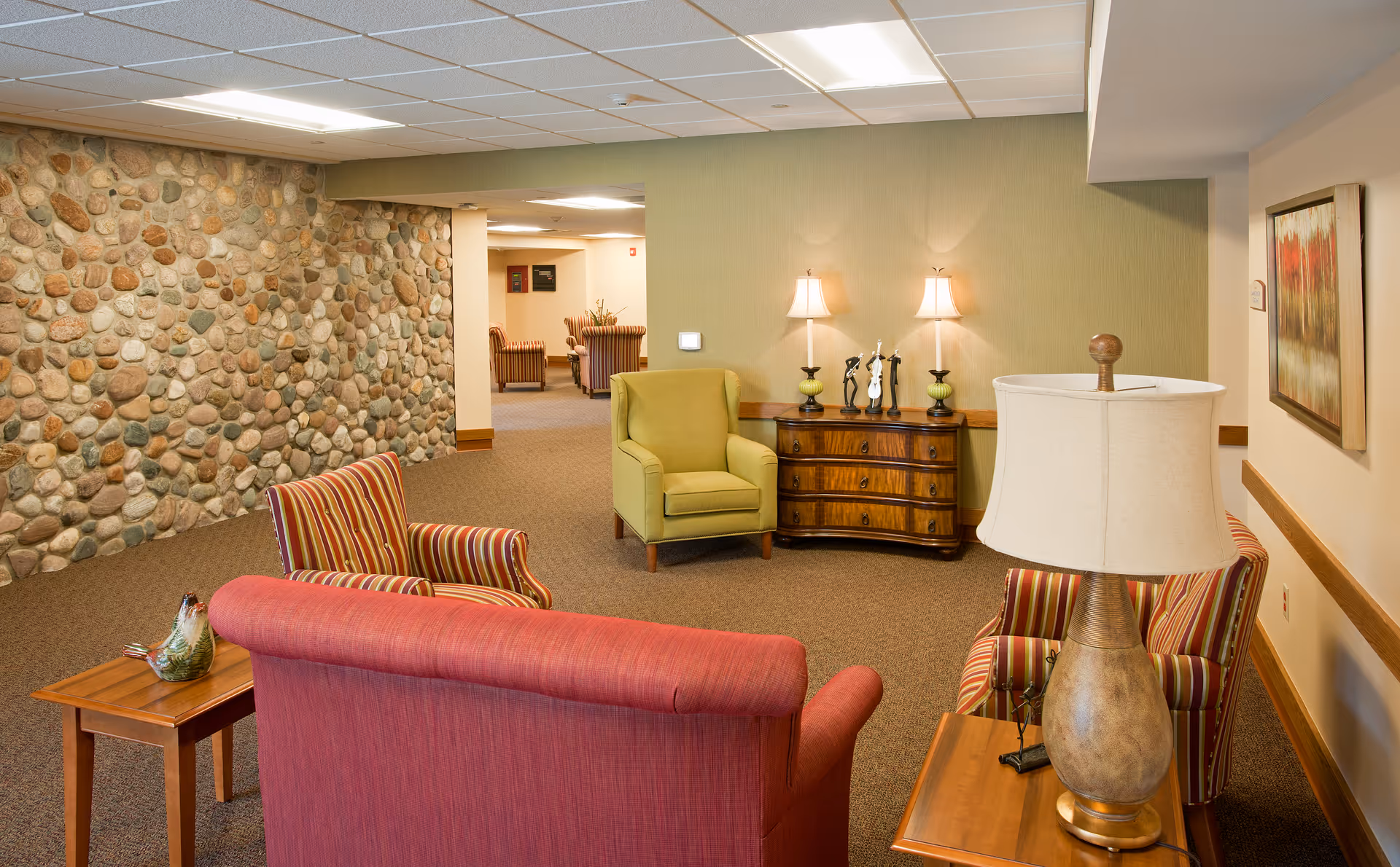 A cozy seating area in a senior care facility with a red sofa, two striped armchairs, a green armchair, wooden side tables with lamps, and a wooden chest of drawers against a green wall. The room features a stone accent wall and carpeted floor, with additional seating visible in the background.