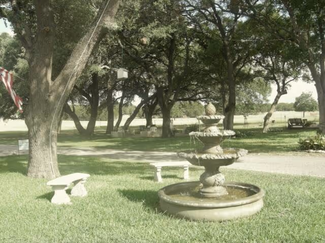A three-tiered stone fountain on a grassy lawn with benches and large oak trees in the background.