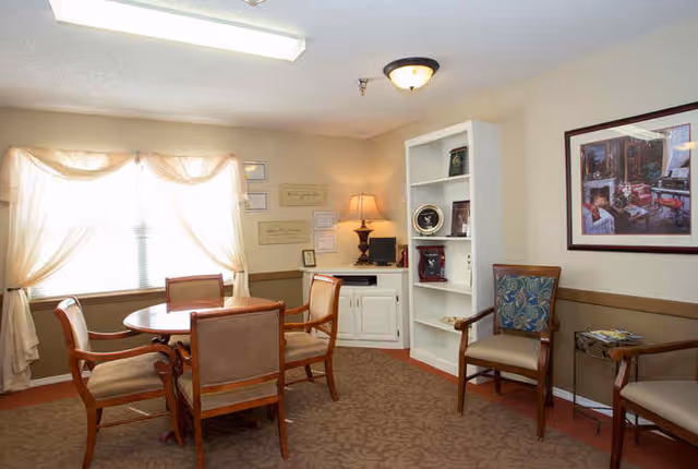 A cozy room with a round wooden table surrounded by four upholstered chairs. There is a window with sheer curtains letting in natural light. Against the wall is a white built-in cabinet with a lamp and computer on top, and a white bookshelf with decorative items. Two additional upholstered chairs are placed near a small side table with magazines. A framed picture hangs on the wall.