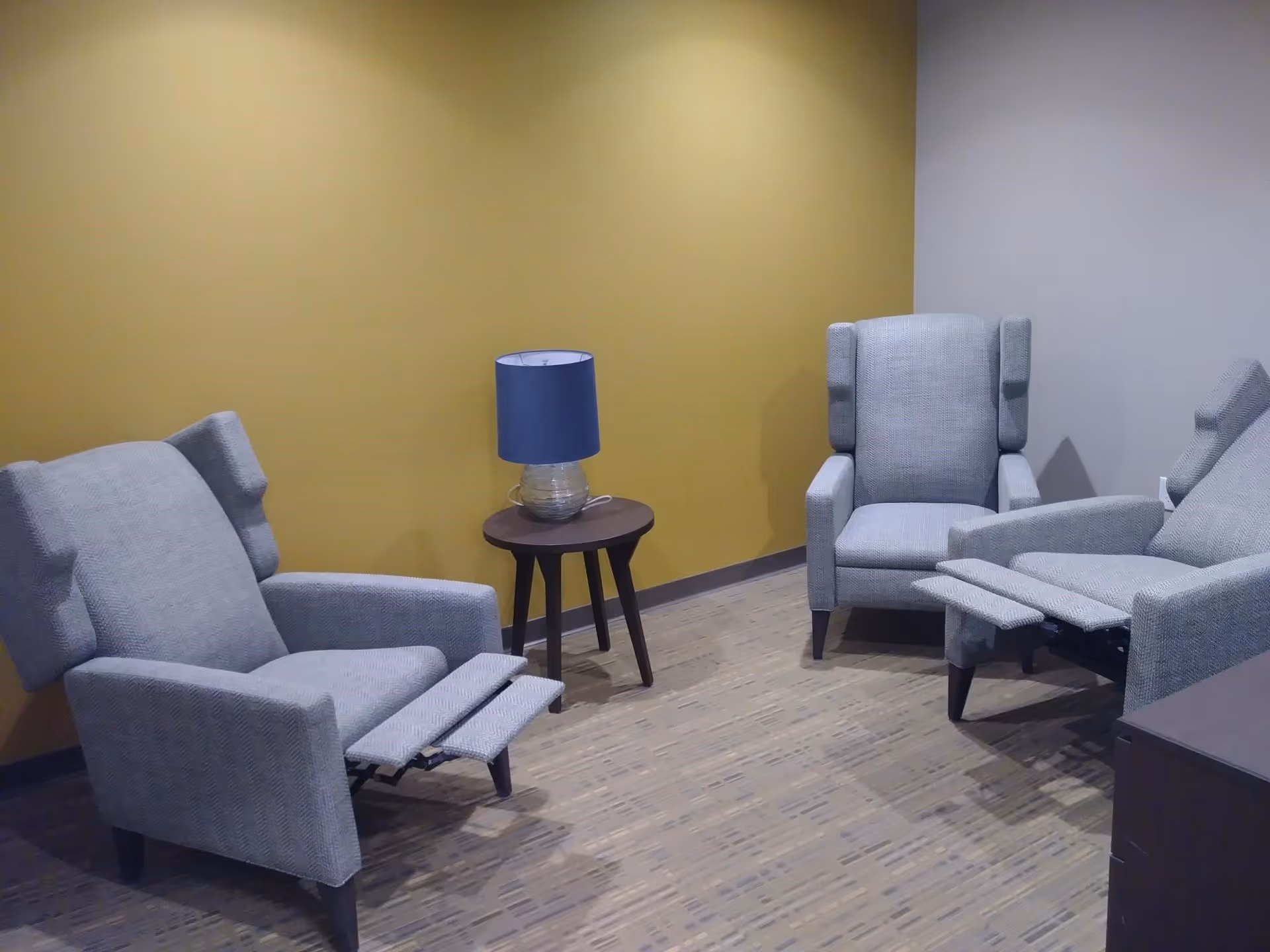 A small seating area with three gray upholstered recliner chairs arranged around a small round wooden side table with a blue lampshade. The walls are painted yellow and light gray, and the floor has a patterned carpet.