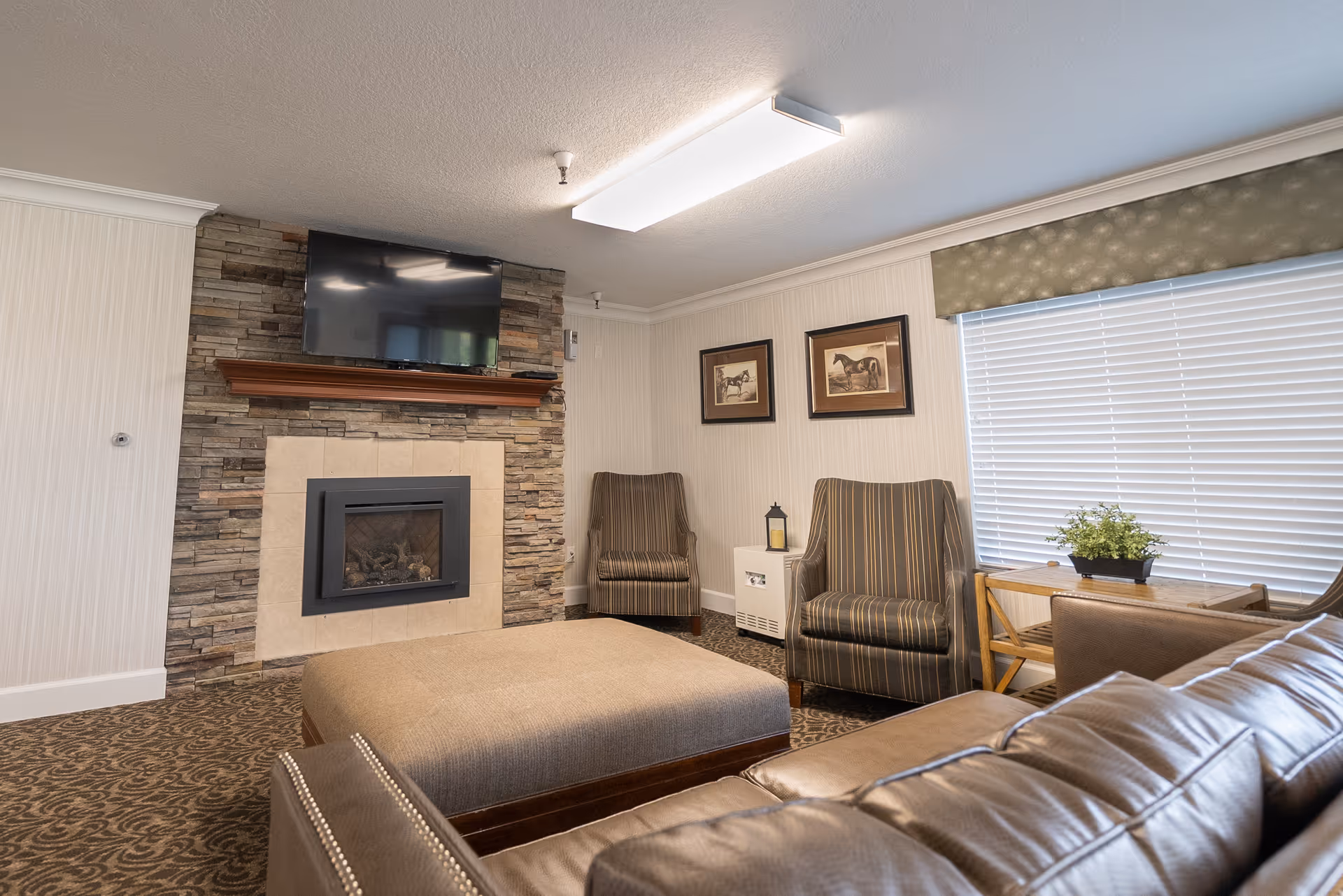 A cozy living room area featuring a stone fireplace with a mounted flat-screen TV above it. The room includes a large ottoman, a leather sofa, two striped armchairs, a small white cabinet with a lantern on top, framed horse artwork on the wall, a window with blinds and a valance, and a small wooden table with a potted plant.