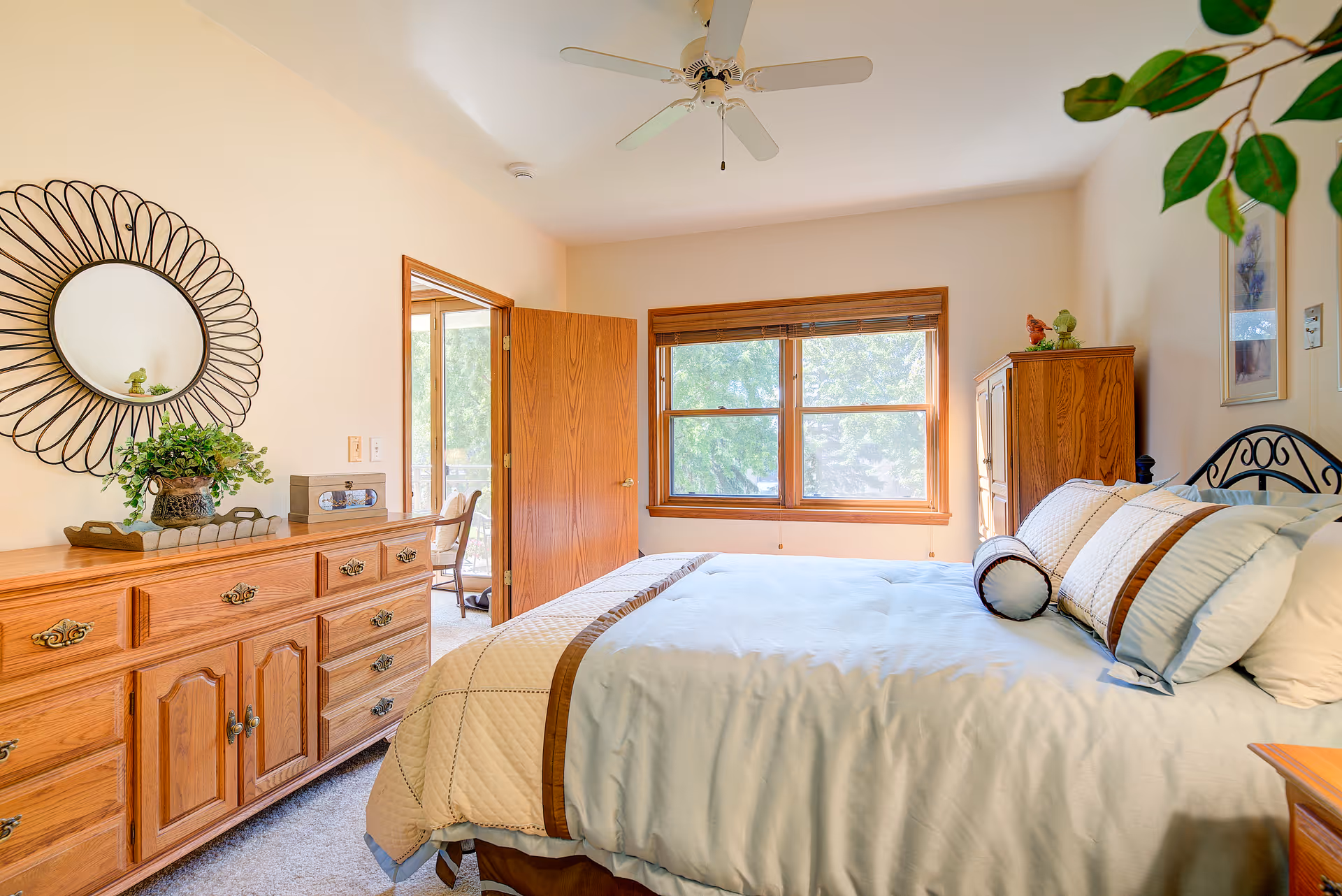 A bright bedroom with a large bed covered in white and brown bedding. There is a wooden dresser with a decorative round mirror above it on the left side, and a wooden cabinet on the right side near a window with wooden blinds. A ceiling fan is mounted on the white ceiling, and a door is open leading to another room with a chair visible.