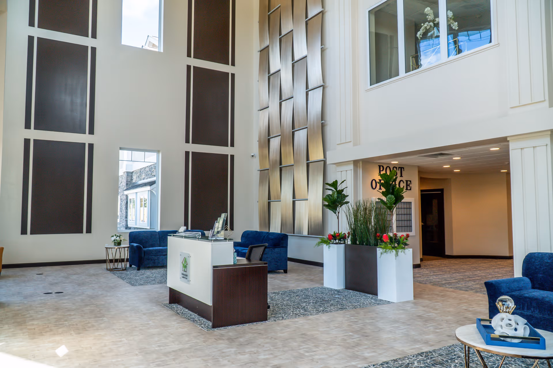 A spacious and modern lobby area with high ceilings and large windows. The space features blue upholstered seating, a reception desk, decorative plants, and a sign indicating the post office area. The walls have tall, dark rectangular panels and a textured metallic wall art installation.
