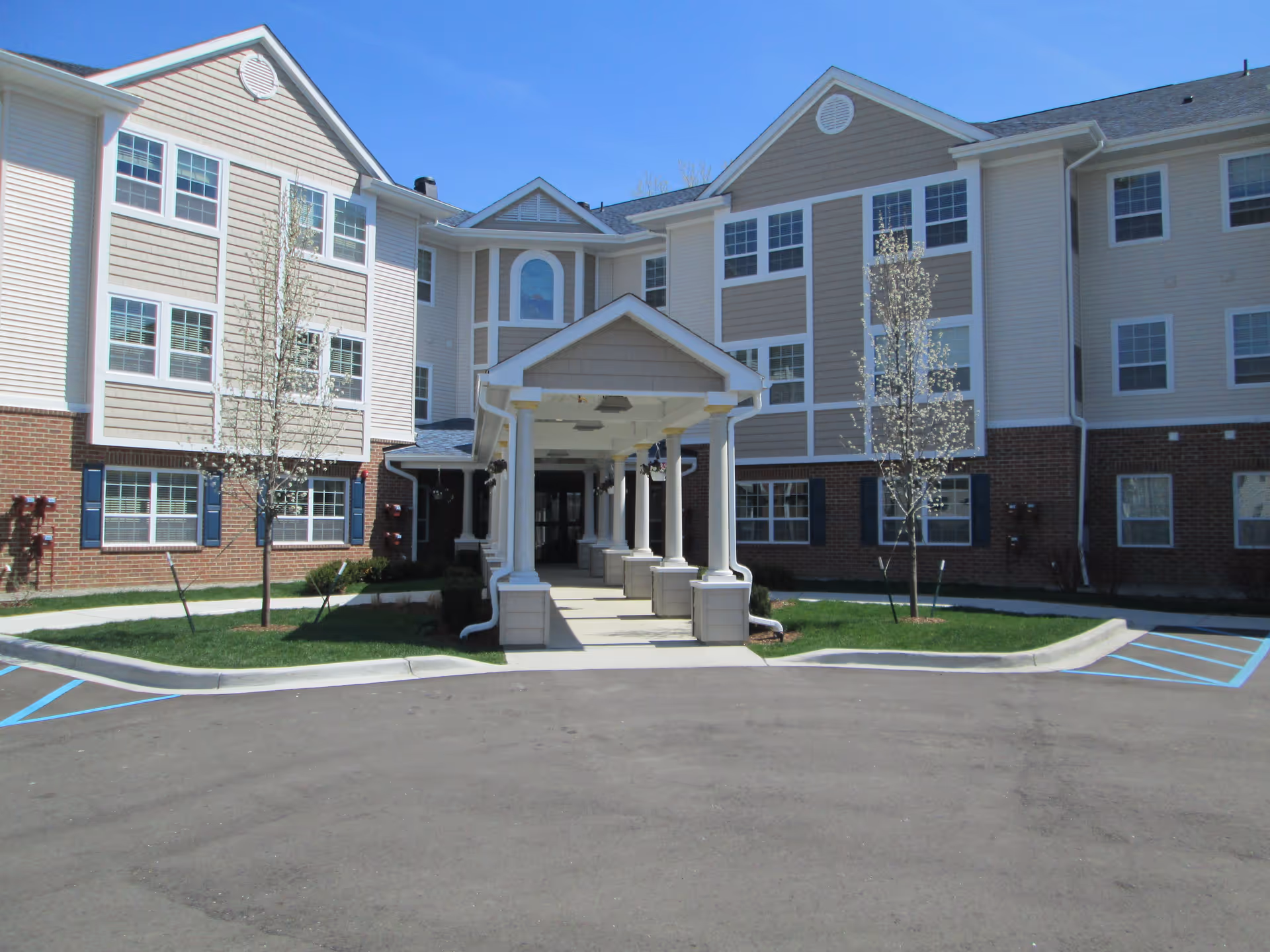 Exterior view of a multi-story senior living facility building with beige siding and brick accents. The entrance features a covered walkway with white columns. There are small trees and green grass on either side of the entrance, and the sky is clear and blue.