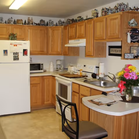 A kitchen with wooden cabinets, a white refrigerator, stove, microwave, coffee maker, and a sink. There is a small counter with a chair, some papers, and a vase with colorful flowers. Decorative miniature houses line the top of the cabinets.