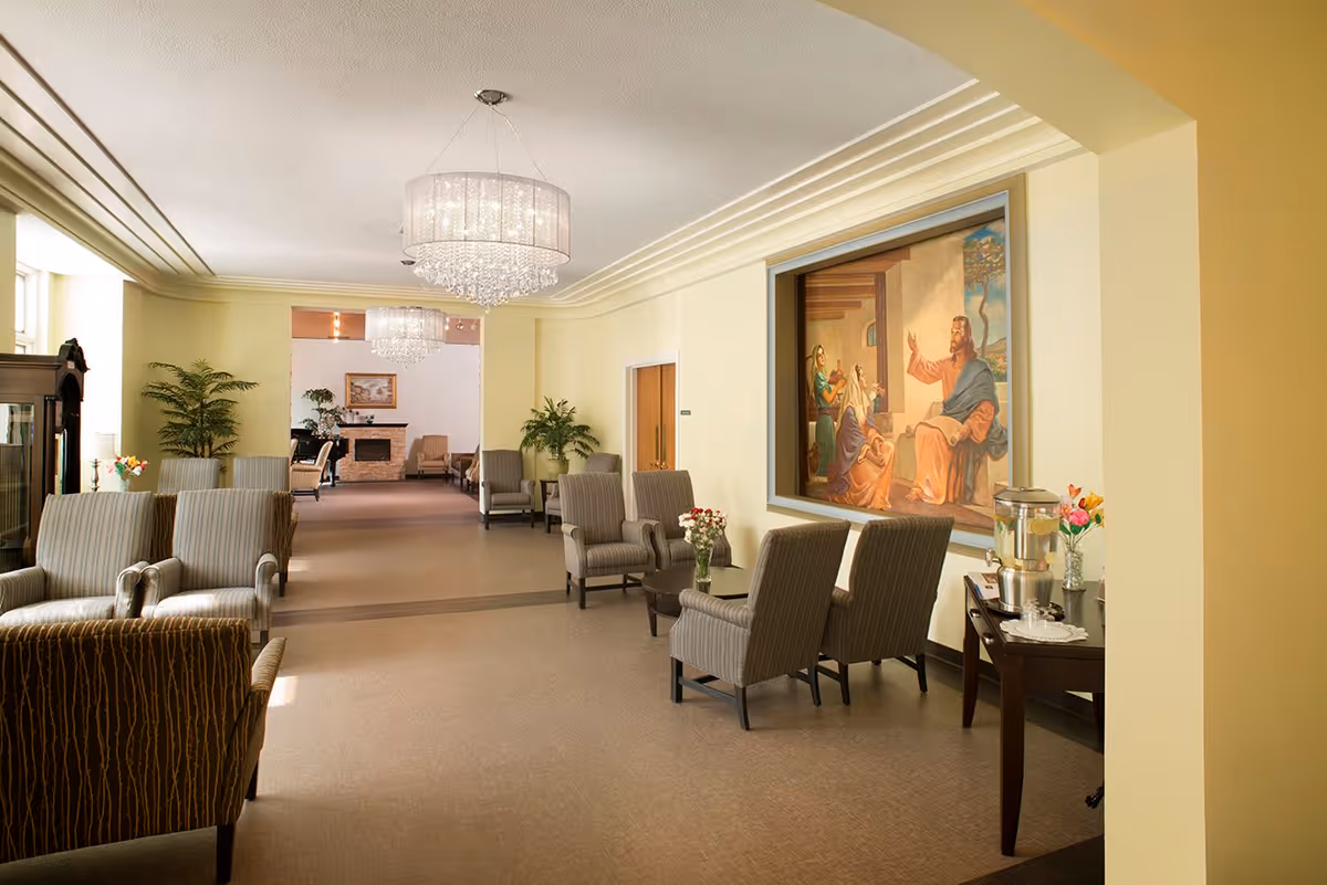 A spacious, well-lit lounge area with multiple striped armchairs arranged along the walls. The room features two large crystal chandeliers hanging from the ceiling, potted plants, and a large framed painting depicting a biblical scene on one wall. A small table with a water dispenser and flowers is visible on the right side. The far end of the room has a fireplace and additional seating.