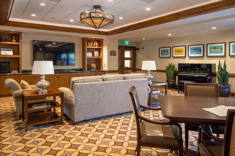 A cozy living room area in The Orchards Health Center featuring a large flat-screen TV mounted on a wooden cabinet wall, a beige sofa with side tables and lamps, a wooden dining table with chairs, a black piano against the wall, potted plants, and framed fish artwork hanging above the piano. The room has patterned flooring and recessed lighting in the ceiling.
