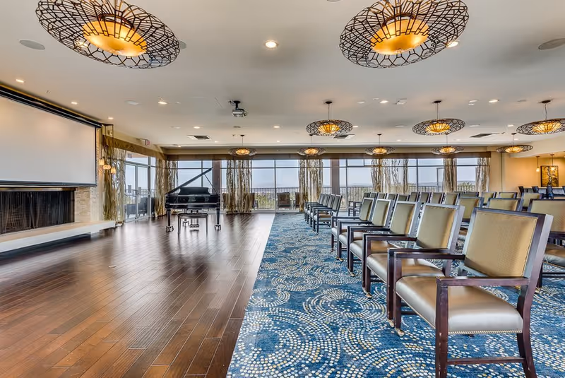 A spacious event room with rows of chairs arranged on a patterned blue carpet. The room features large windows with sheer curtains allowing natural light to fill the space. There is a grand piano near the windows and a large projection screen on the opposite wall. Modern circular ceiling lights illuminate the room.