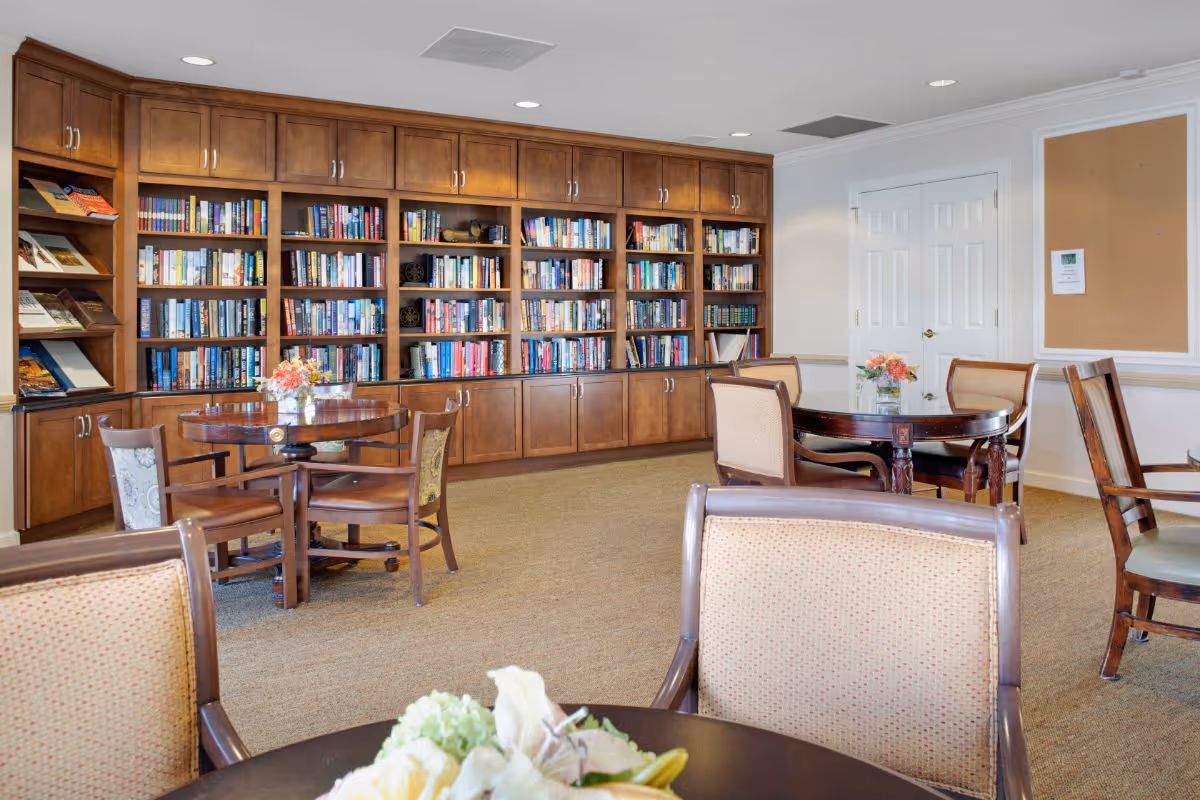 A bright communal reading room with wall-length wooden bookshelves, round tables and upholstered chairs.