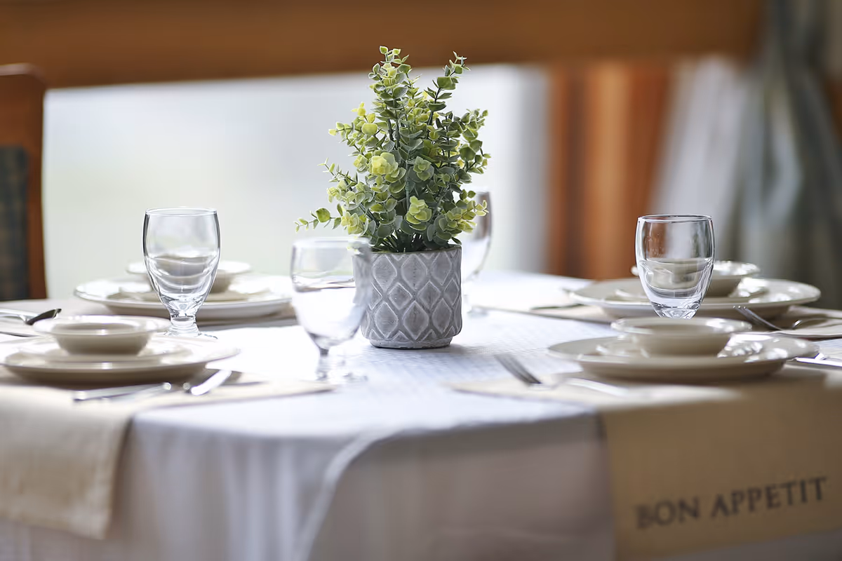 A dining table set with white plates, bowls, silverware, and clear drinking glasses. A small potted plant with green leaves is placed in the center of the table. The table is covered with a white tablecloth and a beige runner with the words 'BON APPETIT' printed on it.