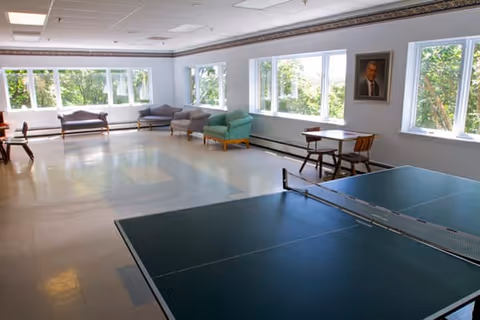 Bright communal activity room with a ping-pong table in the foreground, chairs and sofas along a row of windows, and a portrait on the wall.