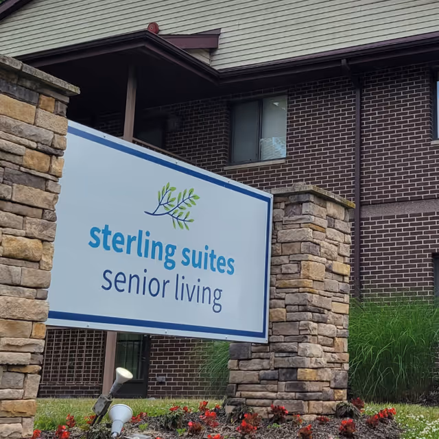Outdoor view of a sign for Sterling Suites Senior Living mounted between two stone pillars with a brick building and greenery in the background.