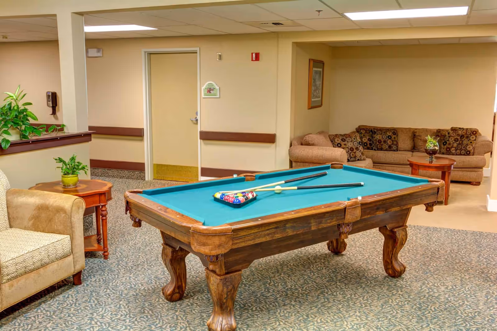 A common room featuring a pool table in the center with sofas, armchairs, and side tables around it.
