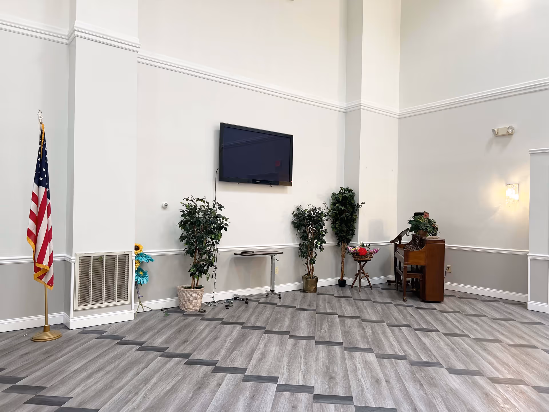 A spacious room with light gray walls and a patterned gray floor. There is an American flag on a stand to the left, a flat-screen TV mounted on the wall, several potted plants, a small table with flowers, and a wooden piano with a plant on top. The room has high ceilings and a wall sconce light fixture.
