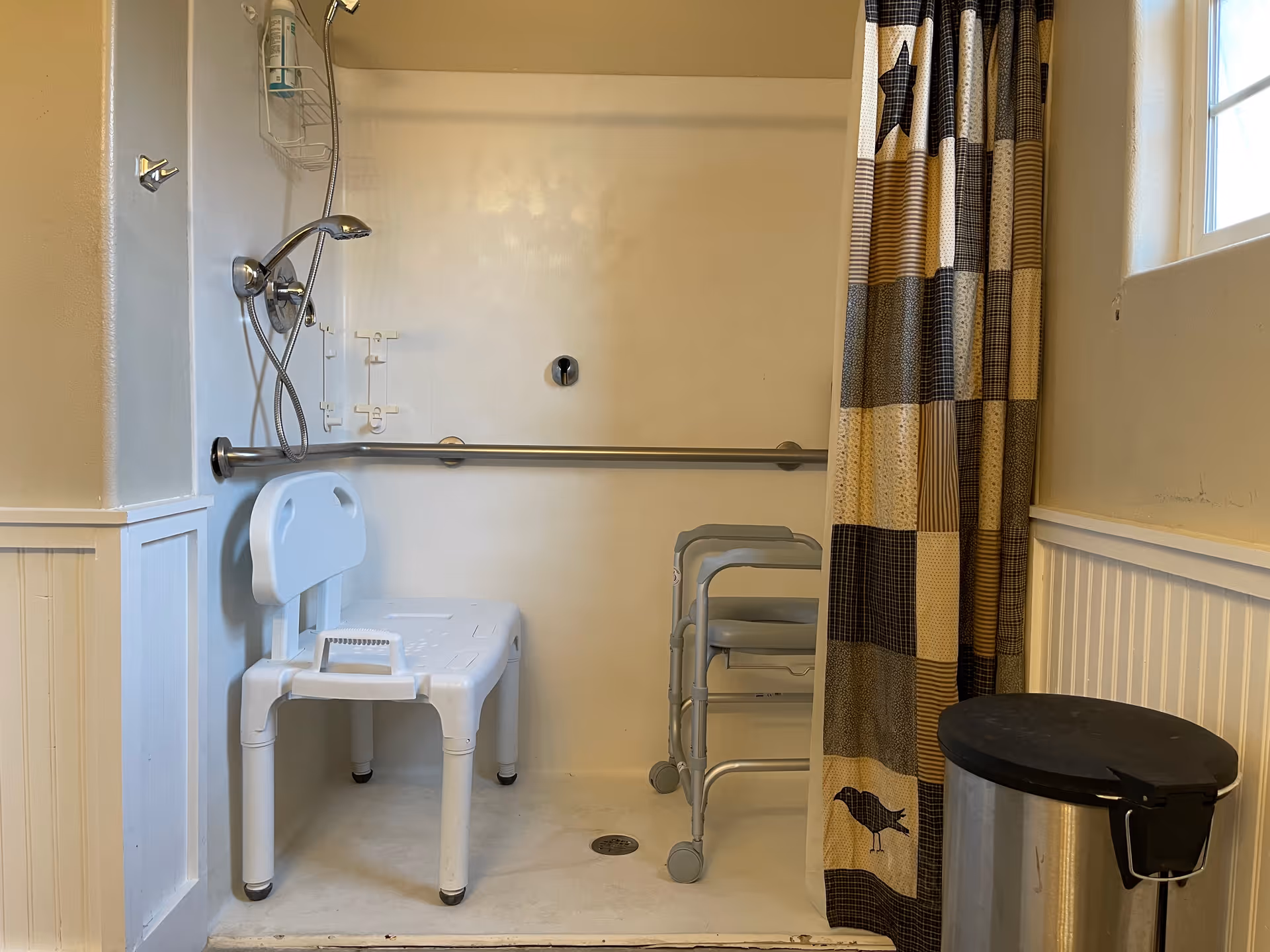 Accessible shower area with a white plastic shower chair, a metal grab bar on the wall, a handheld showerhead, a patterned shower curtain, a small window, and a metal trash can nearby.