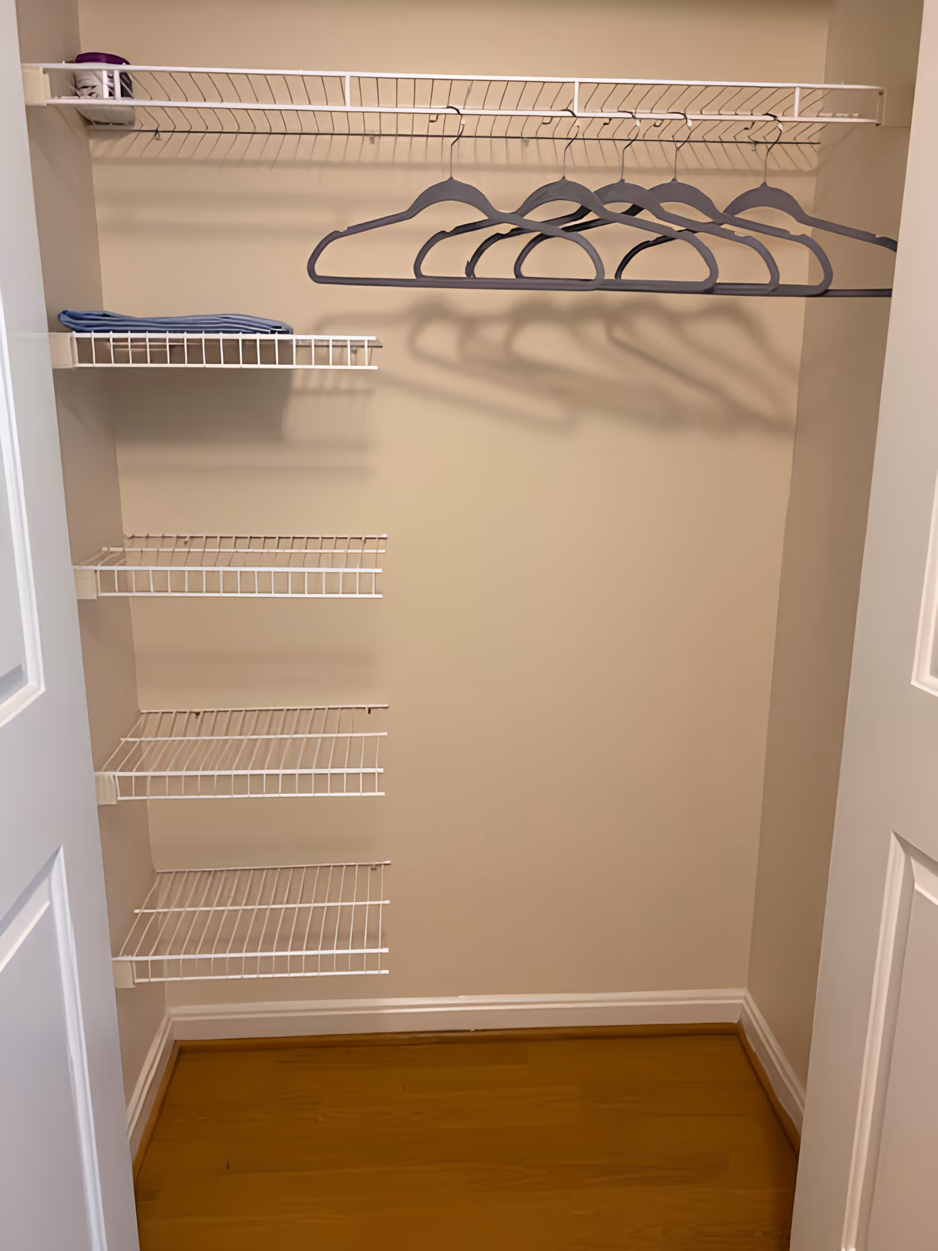 An empty closet with white wire shelves on the left wall and a white wire hanging rack with several empty hangers. The closet has beige walls and a wooden floor.