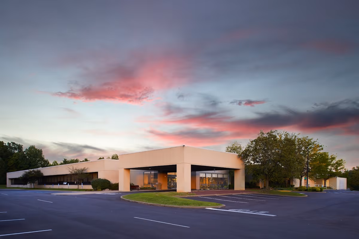Exterior view of Southern Indiana Rehabilitation Hospital - Skilled Nursing Facility at dusk with a partly cloudy sky showing pink and purple hues. The building is a single-story structure with a covered entrance and surrounding parking lot with empty parking spaces. Trees and shrubs are visible around the building.