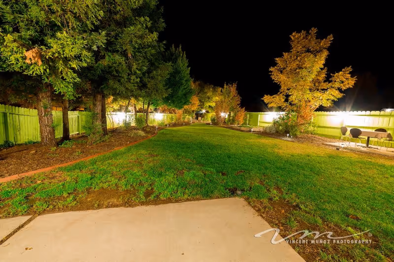 Well-lit fenced backyard at night with a grassy lawn, trees, and a small patio area with chairs.