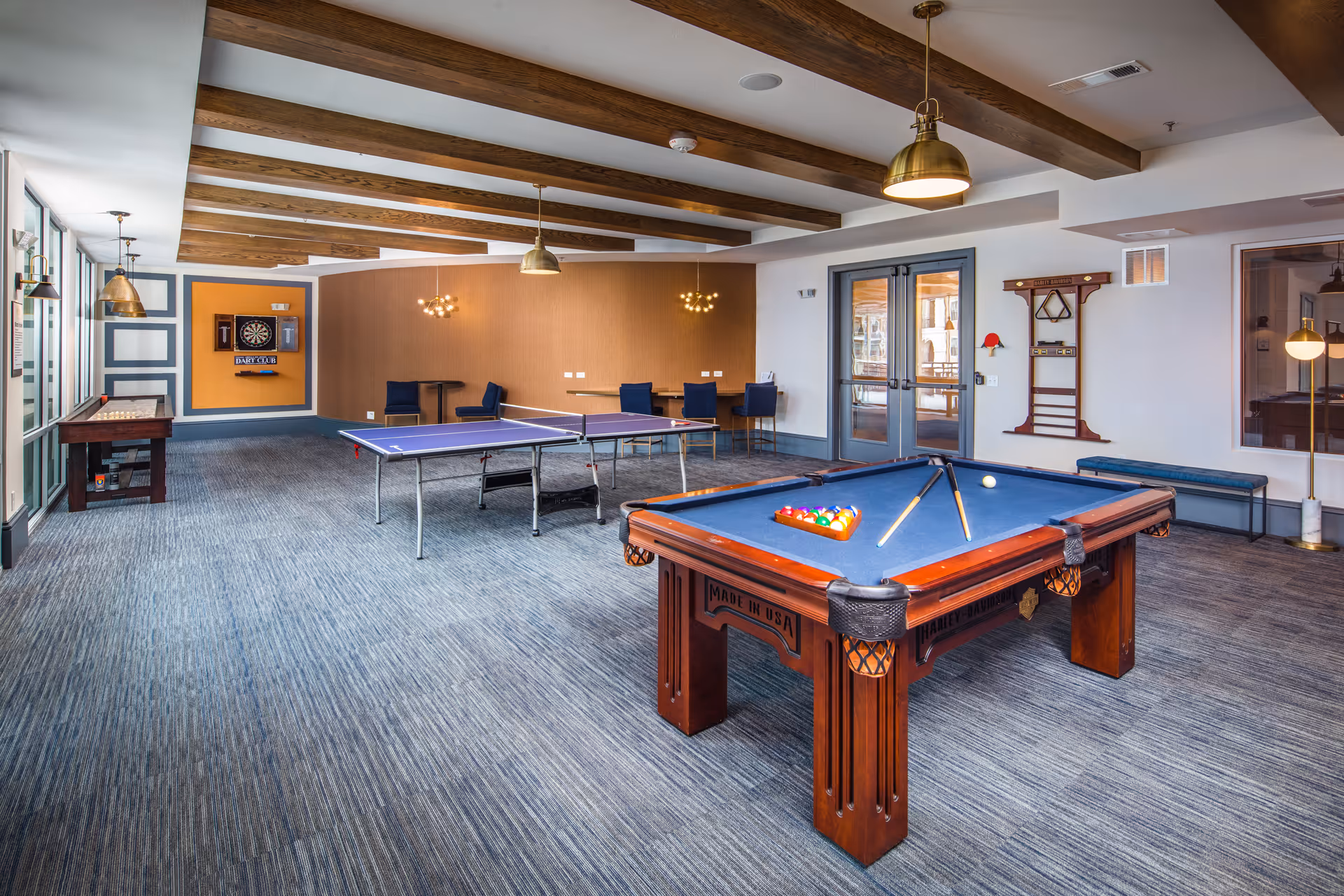 A spacious game room with a pool table, ping pong table, and shuffleboard. The room features wooden ceiling beams, modern pendant lights, and a dartboard mounted on an orange accent wall. There are chairs and small tables along the walls, and double glass doors leading to another area.