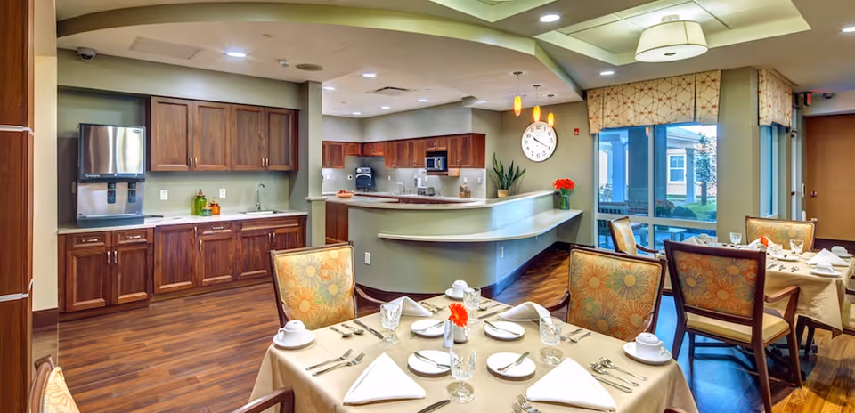 A bright and clean dining area in a senior living facility with tables set for a meal, including plates, glasses, napkins, and silverware. The room features wooden flooring, floral upholstered chairs, a kitchen area with wooden cabinets, a water dispenser, and a large clock on the wall. Large windows provide natural light and a view of the outside.