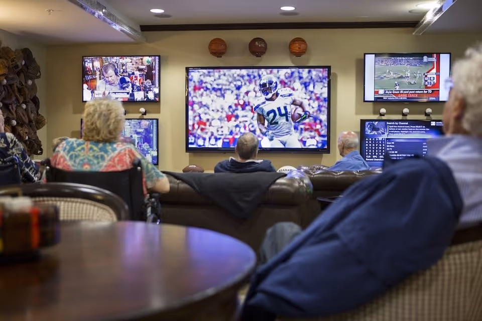 A group of elderly people sitting in a living room area watching multiple television screens mounted on the wall, one showing a football game. The room has comfortable seating including a wheelchair and couches, with sports memorabilia such as basketballs displayed above the TVs.