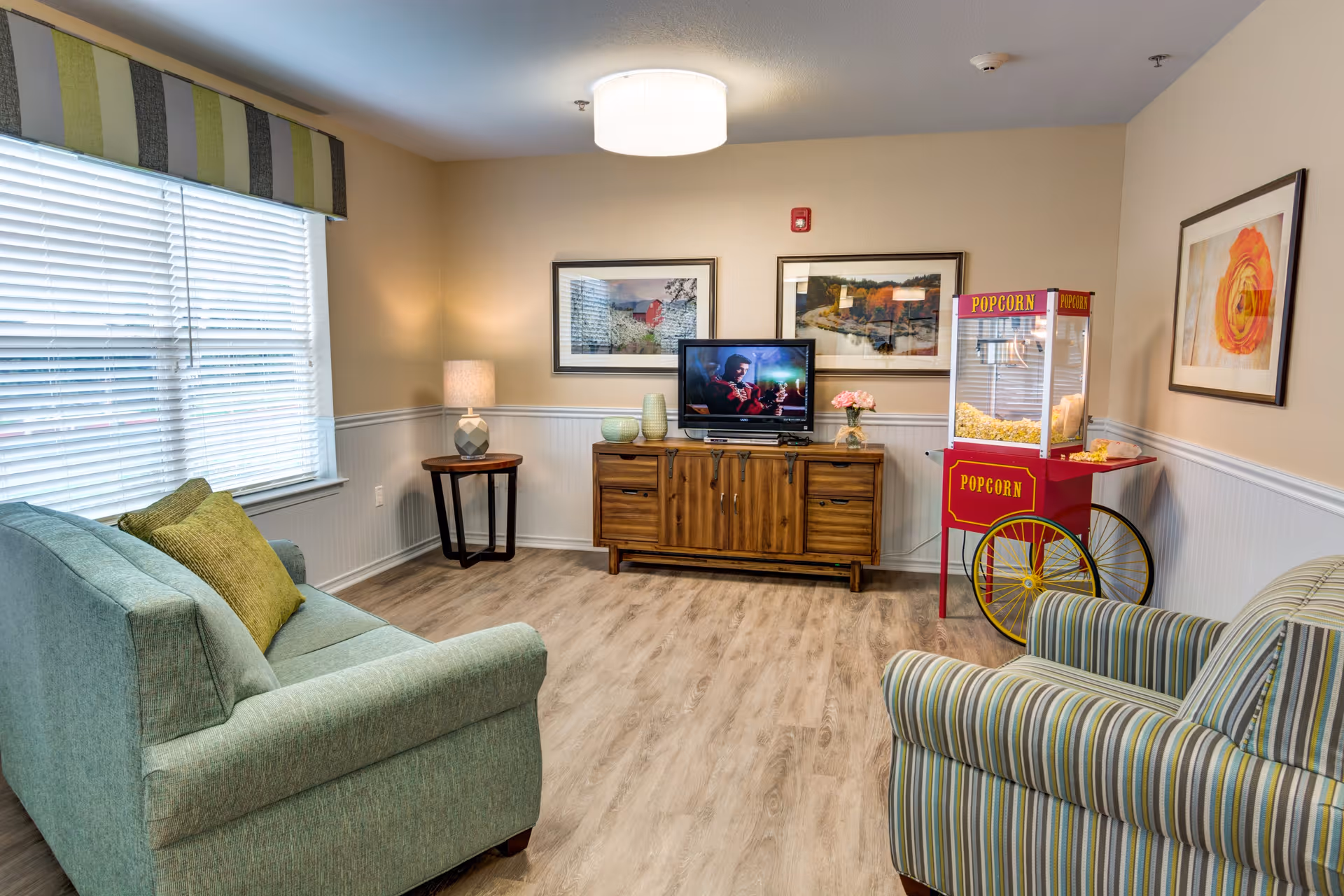 A comfortable common living room with a sofa, striped armchair, TV on a wooden console, and a popcorn machine.