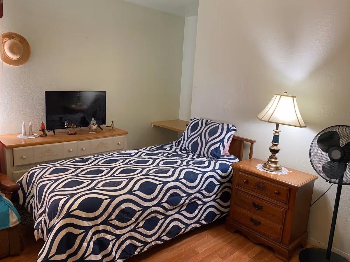 A cozy bedroom with a single bed covered in a blue and white patterned bedspread. Next to the bed is a wooden nightstand with a lit lamp and a standing fan beside it. Across from the bed is a dresser with a flat-screen TV and several small decorative figurines. A straw hat hangs on the wall above the dresser.