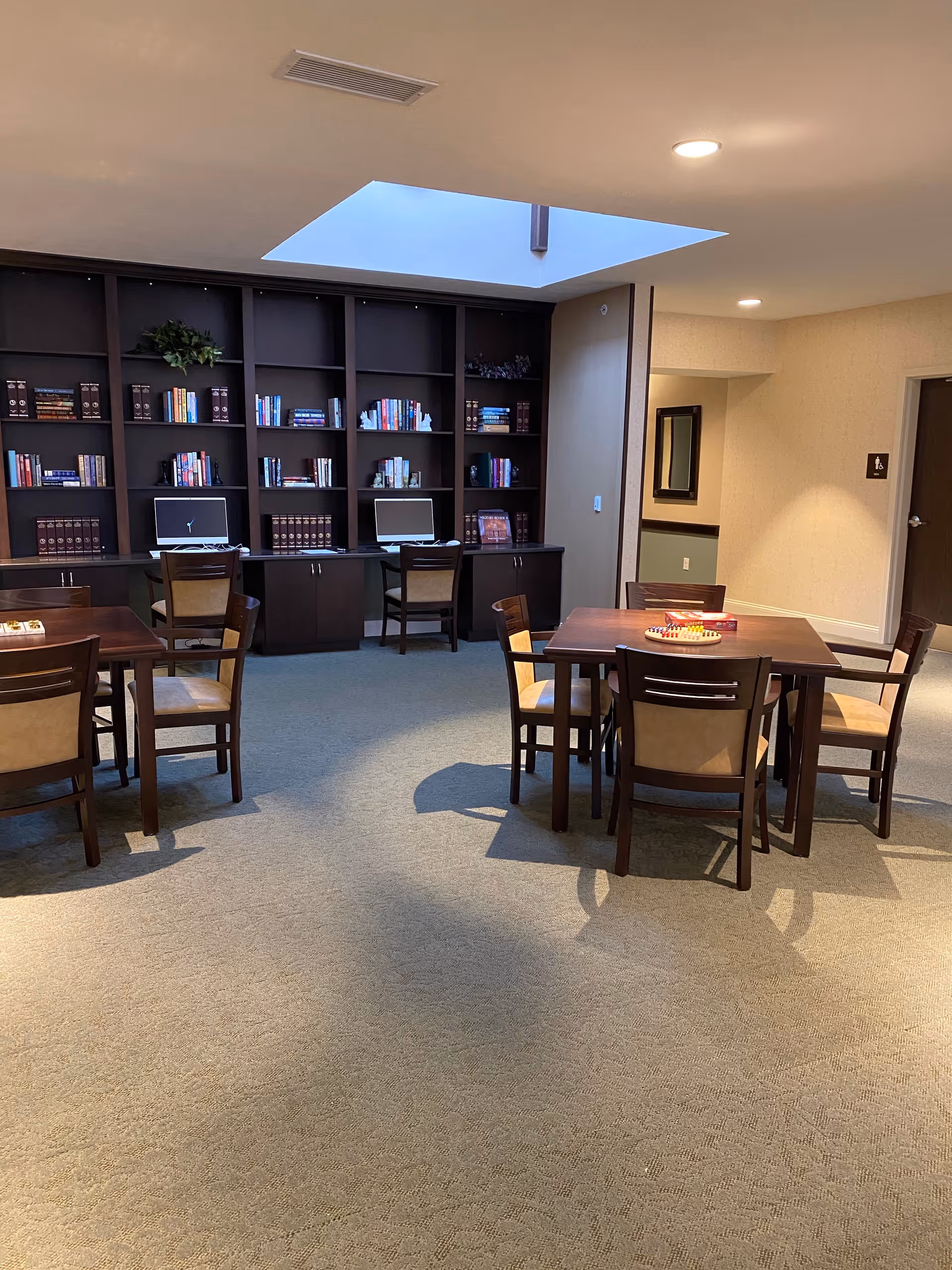A quiet common area with two wooden tables and chairs arranged for seating. In the background, there is a dark wooden bookshelf filled with books and two computer stations with chairs. The room is lit by ceiling lights and a skylight above.