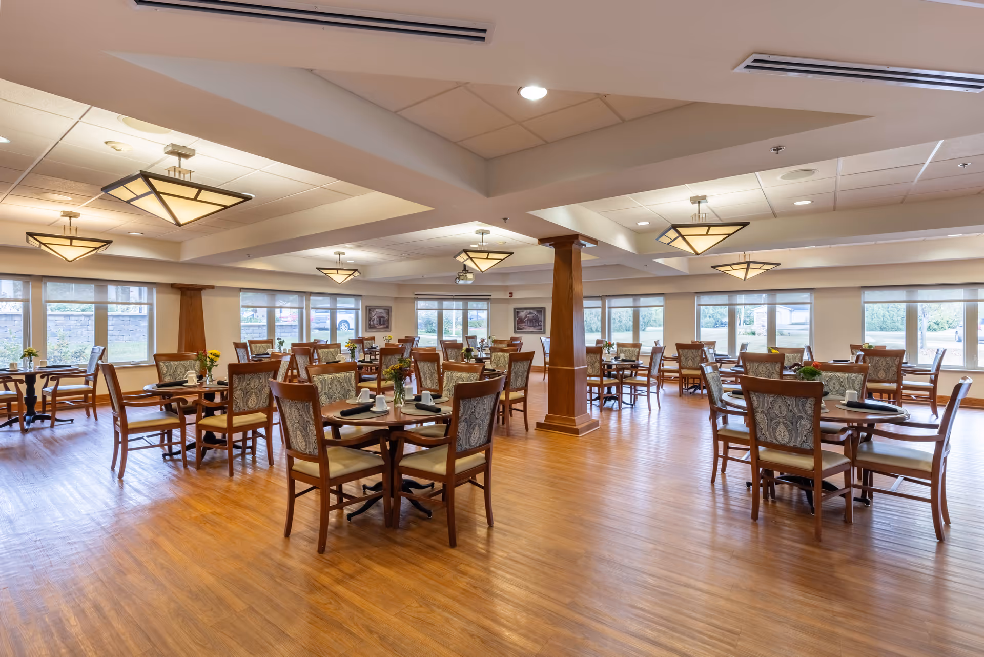 Spacious dining room with multiple wooden tables and chairs arranged neatly. Each table is set with plates, napkins, and small flower arrangements. Large windows line the walls, allowing natural light to fill the room. The ceiling features recessed lighting and decorative hanging light fixtures. The floor is made of polished wood.
