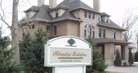 A large, multi-story brick building with a covered entrance, surrounded by trees and shrubs. In front of the building is a white sign with green and gold accents that reads 'Rhodes Estates Assisted Living.'