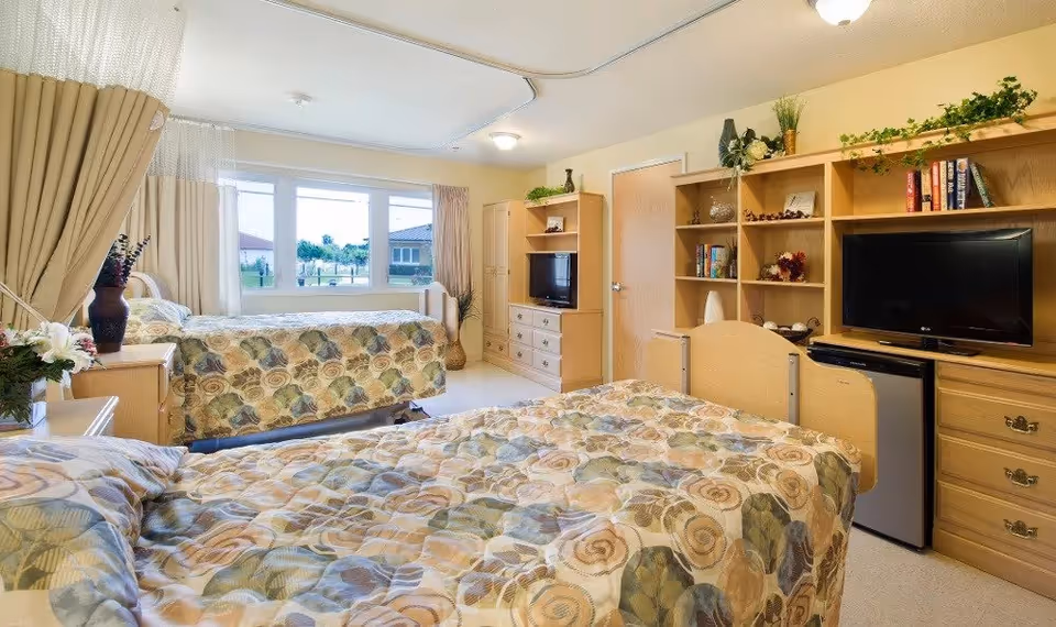A bright and cozy room with two beds covered in patterned bedspreads. The room features large windows with beige curtains letting in natural light. There are wooden cabinets and shelves with decorative items, books, and a television. A small refrigerator is also visible under the shelving unit.