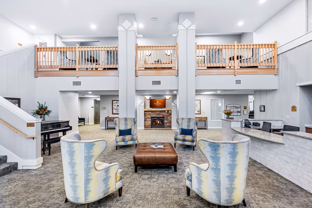A spacious senior living facility common area with four patterned armchairs arranged around a brown leather ottoman. In the background, there is a stone fireplace with a wooden mantel, a piano to the left, and a reception desk to the right. The room features a high ceiling with a wooden balcony railing on the upper level.