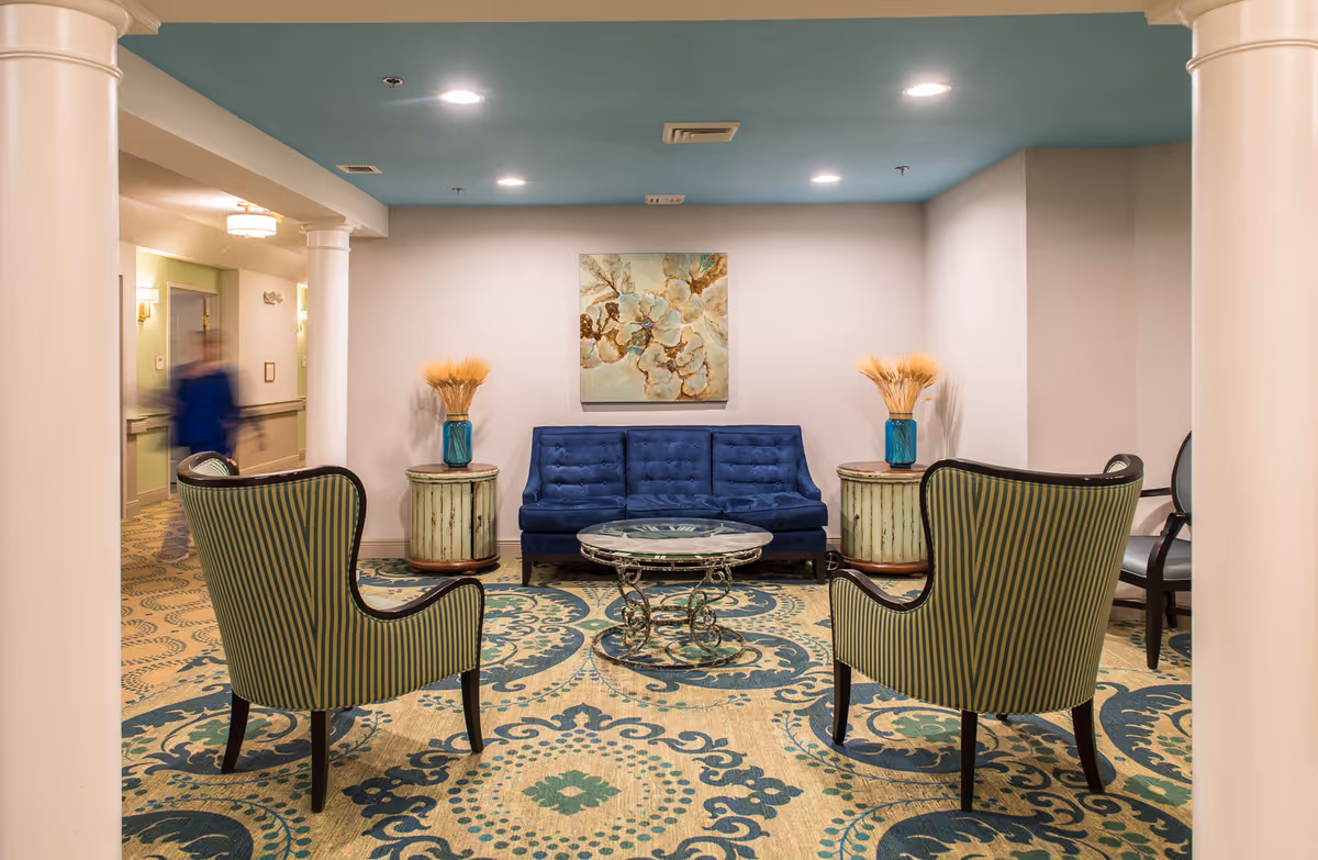 A cozy sitting area in a senior living facility featuring a blue tufted sofa, two striped armchairs, a round glass coffee table with ornate metal legs, and two small round side tables with blue vases holding dried wheat stalks. The room has a patterned carpet with blue and beige tones, white walls, and a ceiling painted in a muted blue color. A decorative painting hangs on the wall above the sofa, and a hallway is visible to the left with a blurred figure walking.