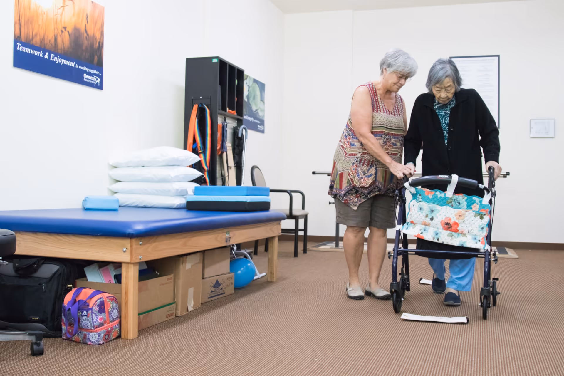 An elderly woman using a walker is assisted by a caregiver in a therapy or rehabilitation room. The room has a padded therapy table with pillows and exercise equipment, and motivational posters on the walls.