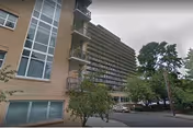 Exterior view of a multi-story senior living facility building with balconies, surrounded by trees and a parking area with several cars.