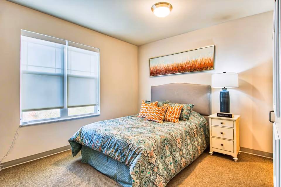 A bedroom with a bed covered in a patterned blue and green bedspread with orange and white decorative pillows. There is a nightstand with three drawers next to the bed, topped with a blue lamp and a book. A large window with white blinds is on the left wall, and a rectangular piece of artwork with orange and white tones hangs above the bed. The room has beige walls and carpeted flooring.