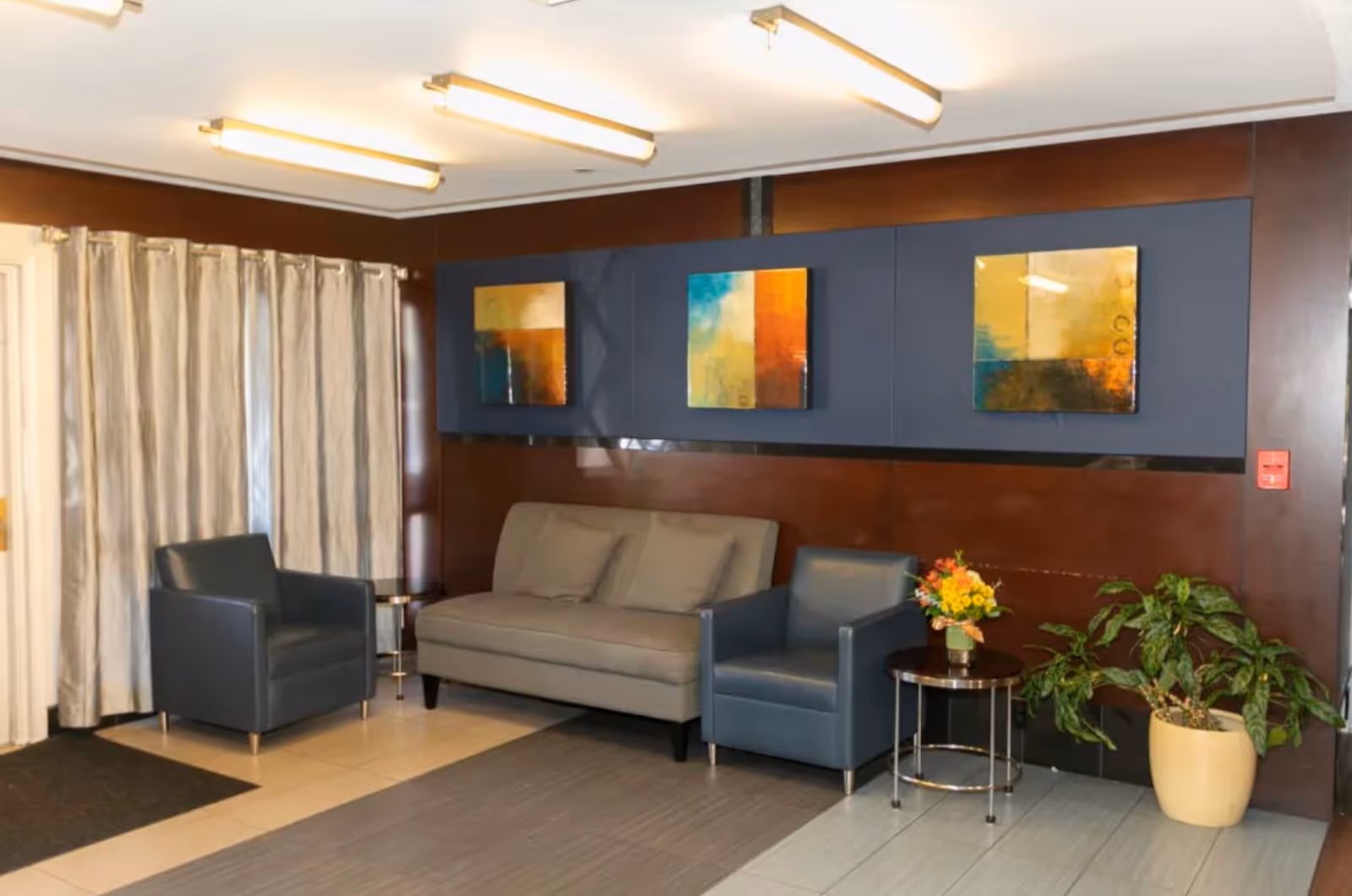 A waiting area with a gray sofa and two dark blue armchairs. There are three abstract paintings on the dark wood-paneled wall behind the sofa. A small round table with a flower arrangement is placed between one armchair and the sofa. A potted plant is on the floor to the right. Beige curtains cover the windows on the left side of the room. The floor has a combination of tiles and a rug, and fluorescent ceiling lights illuminate the space.