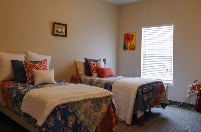 Two twin beds with patterned quilts and decorative pillows in a softly lit assisted living bedroom with a window and wall art.