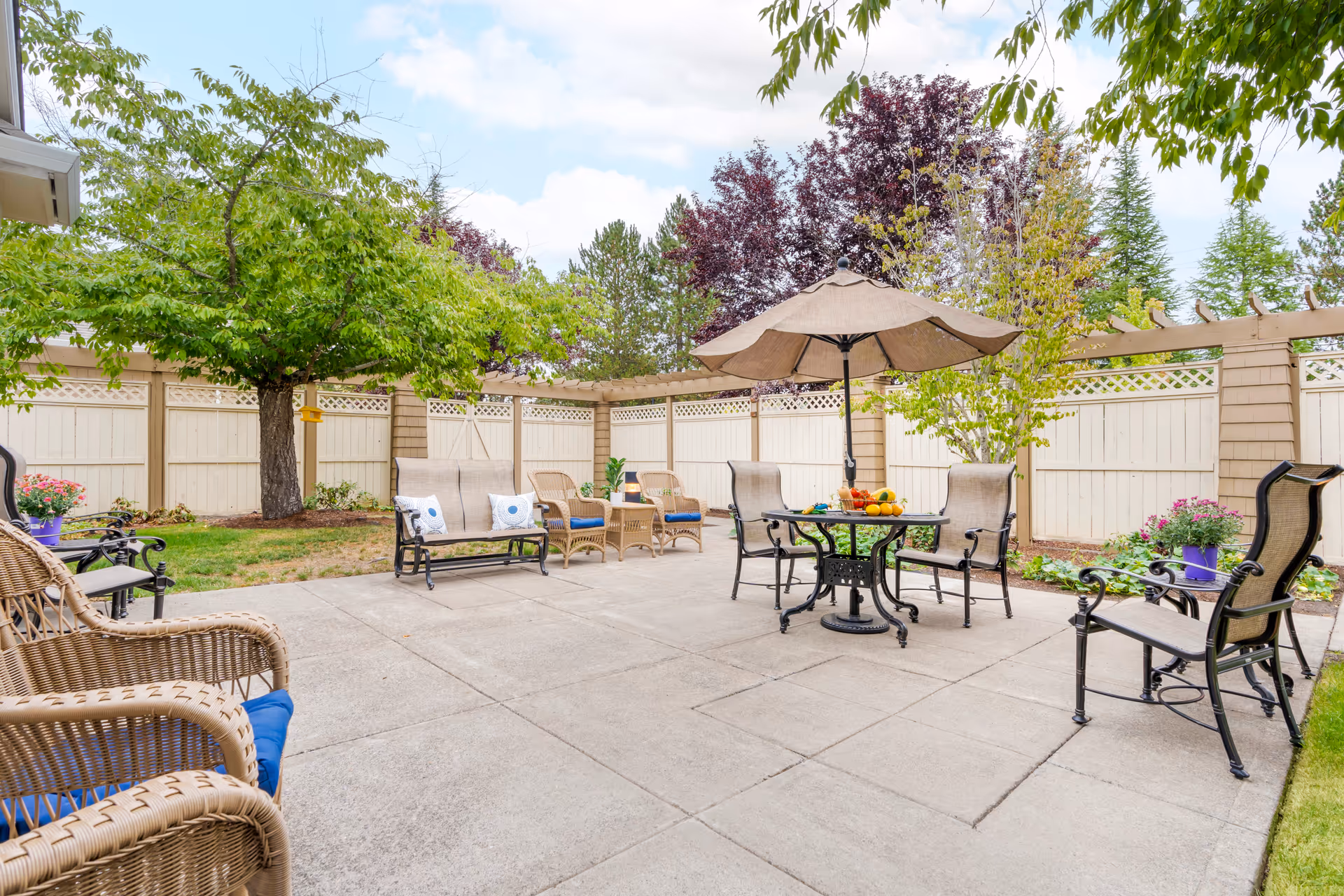 Enclosed sunny patio with a table and umbrella, several chairs and wicker seating on a concrete slab surrounded by a wooden fence and trees.