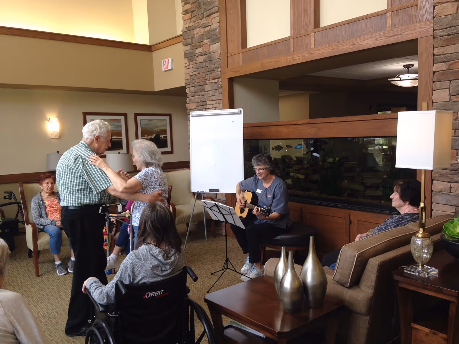A group of elderly people in a senior living facility common area. One elderly man and woman are dancing together while others sit around watching. A woman is playing guitar near a large fish tank. The room is warmly lit with comfortable seating and decorative vases on a table.