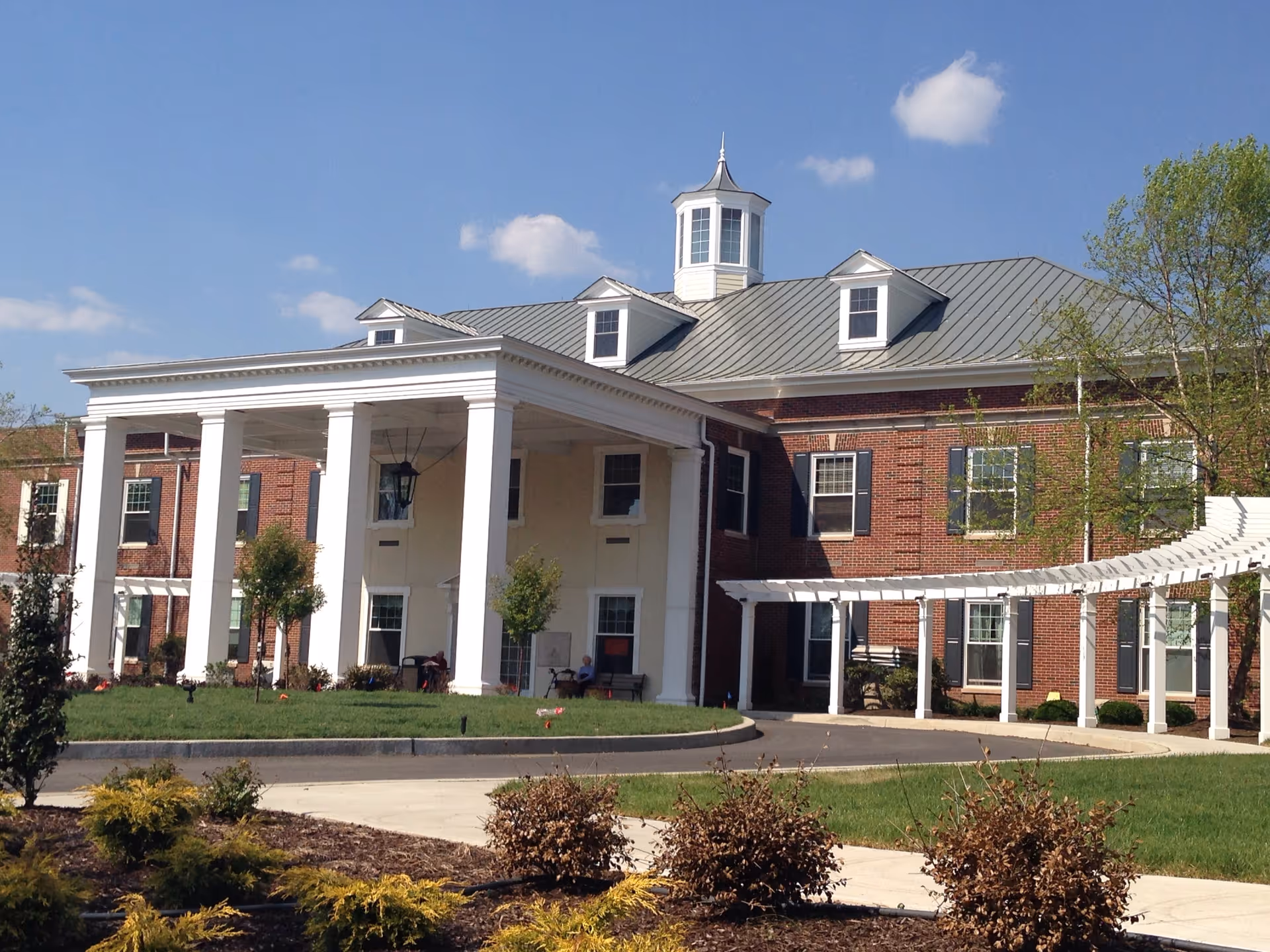 Exterior view of a large brick building with white columns and a covered entrance, surrounded by green grass, shrubs, and trees under a clear blue sky.