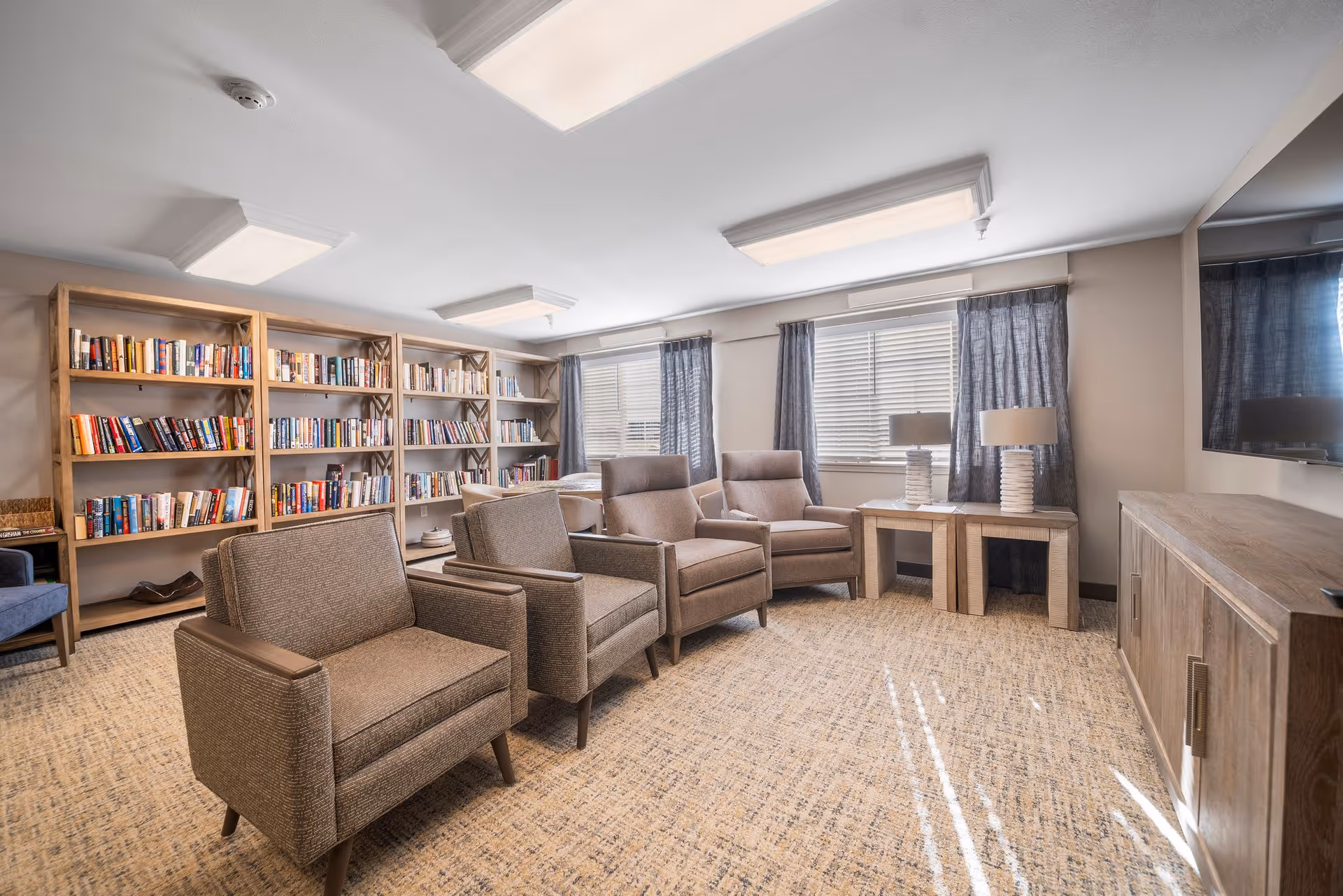 A cozy living room area with four upholstered armchairs arranged in two rows facing a large flat-screen TV mounted on the wall. Behind the chairs, there are large windows with gray curtains allowing natural light to enter. To the left, there are wooden bookshelves filled with books. Two matching side tables with lamps are positioned near the windows. The room has a neutral color palette with beige carpet and light-colored walls.